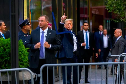 The image captures a scene featuring a group of people outdoors, with a prominent focus on Donald Trump in the center, as suggested by the hint. Trump is dressed in a dark suit with a white shirt, looking assertive as he gestures upwards with his right index finger. The scene is bustling with several individuals, likely security personnel, some of whom are dressed in suits with earpieces, indicating a high-security environment. There is also a uniformed officer present, wearing a navy cap and standing nearby, which further emphasizes the security aspect.
The background includes a reflective glass building, suggesting an urban setting, possibly near an entrance or exit, with metal barriers in the foreground marking a restricted area. The lighting is natural, indicative of a daytime setting, and the overall mood conveys a sense of movement and urgency, typical in public appearances involving high-profile figures. The composition centers around Trump's gesture, making it the focal point amidst the bustling surroundings. The presence of neatly trimmed greenery adds a touch of nature to the otherwise urban scene.