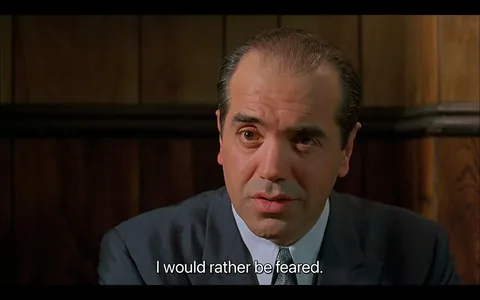 In this frame from "A Bronx Tale," a character is having a serious conversation indoors. The setting features wood-paneled walls, creating an intimate and somewhat somber atmosphere. The character is dressed in a formal suit, suggesting a significant or authoritative role. His expression is intense and focused, reflecting the gravity of the statement, "I would rather be feared," which is displayed as a subtitle. The lighting is soft and natural, emphasizing the character's contemplative demeanor. This scene captures a pivotal moment in the film, emphasizing themes of power and respect, central to the narrative of "A Bronx Tale."
- Captions / subtitles: "I would rather be feared."