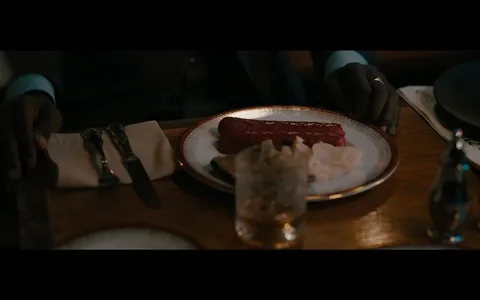 The image is a still from the film "The Menu." It depicts a dining scene, focusing on a plate of food in front of a person. The plate is elegantly set with a piece of meat, likely sausage, garnished with a sauce, and accompanied by what appears to be thin chips or crisps. The setting is a restaurant, with the table neatly arranged with silverware, a cloth napkin, and a glass containing a beverage, possibly whiskey or another brown liquor. The lighting is dim and atmospheric, suggesting a formal dining experience. The person's hands, adorned with a ring, rest on either side of the plate, adding a sense of anticipation or contemplation about the meal. The overall color palette is warm, with rich browns and golds, contributing to the luxurious and slightly mysterious ambiance typical of scenes in "The Menu."