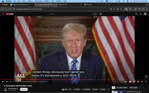 The image is a screenshot from a YouTube video titled "In conversation with President Trump" on the All-In Podcast channel. The scene captures Trump in an indoor setting, likely during an interview or discussion. Two American flags are prominently displayed in the background, framing Trump's image, which emphasizes the political context. Trump is wearing a dark blue suit with a white shirt and a blue-striped tie. He appears to be speaking, with a serious and focused expression. The lighting is bright and consistent, highlighting his features and creating a formal atmosphere typical of political interviews.
The video frame includes subtitles at the bottom, partially reading: "certain things obviously but I gener you know it's transparency and I think." The All-In Podcast logo is visible on the lower left of the screen. This visual setup, along with the presence of the American flags, suggests a formal discussion or address, possibly focusing on topics related to political transparency or governance. The composition centers on Trump, conveying a sense of authority and direct communication to the audience.
