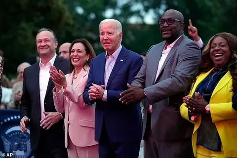 ```
The image captures a group of five people standing outdoors, seemingly at an event. In the center is a man, identified by the hint as Biden, wearing a dark blue suit with a white shirt and a striped tie. To his left, a woman identified as Kamala, is dressed in a light pink suit. Both appear engaged and smiling, suggesting a positive and celebratory atmosphere.
To Biden's right, a man in a dark suit and another individual in a yellow jacket are also present, clapping and smiling, indicating a shared moment of approval or enjoyment. The background shows a blurred crowd, reinforcing the public nature of the gathering. The lighting is warm and soft, likely from the evening or event lighting, creating an inviting ambience. The image suggests a sense of camaraderie and success, possibly at a political or social event.