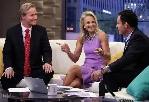 In this image, three people are seated on a talk show set, having a lively conversation. Elisabeth Hasselbeck is prominently featured in the middle, wearing a purple dress and gesturing animatedly with her hands towards the person on her right. She appears engaged and smiling, contributing to a friendly and dynamic atmosphere. The man to her left, dressed in a dark suit and red tie, is looking at her with a smile, suggesting he is listening attentively. The man on her right, also in a suit with a patterned tie, is leaning slightly forward, indicating interest in the conversation. The studio setting includes a modern, cozy setup with a couch and a glass table that has a laptop and a mug on it. The background shows large windows with a view of a cityscape. The lighting is bright and professional, typical for a television studio environment, enhancing the clarity and vibrancy of the scene.