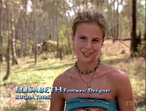 Elisabeth Hasselbeck is shown in an outdoor setting, likely part of a reality TV interview segment. She stands in a wooded area, with trees and some underbrush in the background, suggesting a natural, possibly remote location. The lighting is natural daylight, casting clear, soft shadows. Elisabeth has her hair tied up and is wearing a colorful strapless top along with a beaded necklace. Her expression is friendly and engaging, as she appears to be speaking to the camera. The on-screen text identifies her as "ELISABETH Footwear Designer" from the "Kucha Tribe," indicating her participation in the show "Survivor." The text is positioned in the lower-left corner, with her name in large blue letters, followed by her occupation and tribe affiliation in smaller text. The composition centers on Elisabeth, creating an intimate, personal interview vibe typical of reality television.
- Title: "..."
- Channel / profile: "..."
- Site / app: "..."