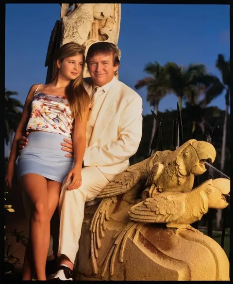 Ivanka and Trump are pictured outdoors in this portrait. Ivanka is seated on a stone sculpture featuring intricately carved parrots. She is wearing a sleeveless floral top and a light blue skirt, her long hair cascading over her shoulders. Trump is beside her, dressed in a crisp white suit, exuding a formal yet relaxed demeanor. The background features lush palm trees and clear skies, indicating a sunny day, enhancing the warm and vibrant ambiance of the scene. The lighting is soft, casting a pleasant glow on their faces. The sculpture adds an element of artistic detail, hinting at an opulent setting. Both appear poised and content, with a serene atmosphere enveloping the scene.