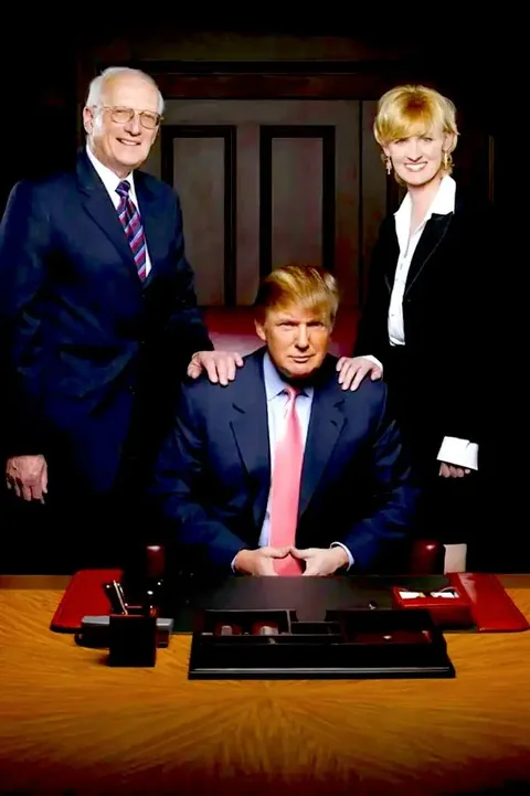 The image depicts a professional setting with three individuals captured in a posed photograph. Donald Trump is seated at a desk in the center, wearing a dark suit with a pink tie. His posture is upright, and his hands are placed together on the desk. To his left, a man is standing, dressed in a suit and tie, with a pleasant expression. To Trump's right, a woman stands wearing a black blazer and white blouse, smiling at the camera.
The background features a wooden panel, giving the setting a formal and authoritative atmosphere. The desk is organized with a leather desk pad, a pen holder, and some desk accessories, enhancing the professional ambience. The lighting is focused, highlighting the individuals and creating a sharp contrast with the darker background, adding to the seriousness of the setting.
