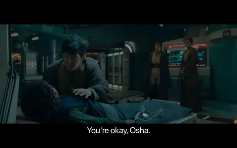 The image depicts a scene set in a futuristic medical bay. In the foreground, a person with short dark hair, dressed in a robe-like outfit, is attending to someone lying on a medical table, referred to as "Osha" by the subtitle. Osha appears to be resting with medical equipment connected to them. The setting is illuminated with soft, bluish lighting, adding to the sci-fi ambiance. In the background, two individuals stand near a display screen, which shows complex graphics, possibly related to medical monitoring. They are also wearing similar robe-like attire. The room has a sleek, metallic design with high-tech elements, reinforcing the sci-fi theme. The overall atmosphere suggests concern and care within a technologically advanced environment.