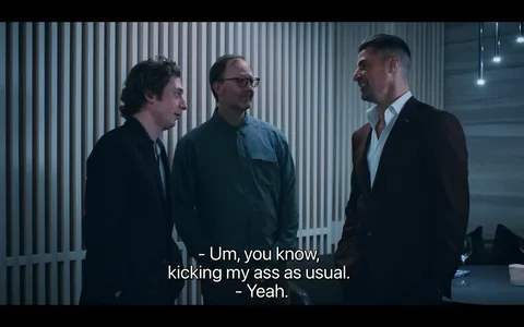 The image shows three men engaged in conversation in a modern, indoor setting, possibly in a dining or social area given the presence of wine glasses on a nearby table. The man on the left is wearing a dark suit with a tie, while the man in the middle has glasses and a button-up shirt. The man on the right is dressed in a dark suit with an open-collared white shirt, giving a casual yet sophisticated appearance. The background features a stylish, vertical striped wall, and the lighting is soft, suggesting an evening or intimate setting. The mood appears relaxed and informal, as indicated by the casual posture and subtle smiles of the men. The subtitles at the bottom of the image read, "- Um, you know, kicking my ass as usual. - Yeah." This exchange suggests a lighthearted or humorous conversation.