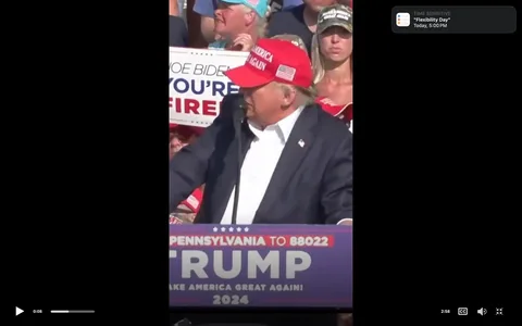 Donald Trump is depicted at a political rally, standing at a podium. He is wearing a red "Make America Great Again" cap and a dark suit with a white shirt. The podium features text that includes "PENNSYLVANIA TO 88022," "TRUMP," and "MAKE AMERICA GREAT AGAIN! 2024." The rally setting is outdoors, with a noticeable crowd in the background. Supporters are holding signs, one reading "JOE BIDEN YOU'RE FIRED." The atmosphere appears energetic, with a mix of casual and patriotic attire among the crowd. A notification titled "TIME SENSITIVE" for "Flexibility Day" appears on the top right, indicating a digital screen capture. The lighting suggests a sunny day, enhancing the vividness of the red and blue colors associated with the event.