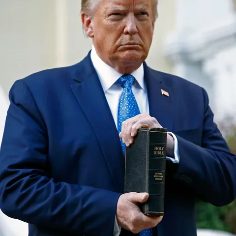 Donald Trump is depicted in a formal outdoor setting, holding a copy of the "Holy Bible" in his right hand. He is dressed in a dark blue suit with a light blue patterned tie, projecting a serious and focused expression. The Bible, a "Revised Standard Version," is prominently displayed with its title visible in gold lettering against the black cover. The background is slightly blurred, suggesting it is an external location, possibly near a building, enhancing the focus on Donald Trump and the Bible. His posture is upright, conveying a sense of gravitas and formality. The lighting is natural, emphasizing the outdoor setting, and the colors are vivid, highlighting the contrast between the dark suit and the lighter background. This image captures a moment of symbolic significance, possibly intended to convey a message of authority and tradition.