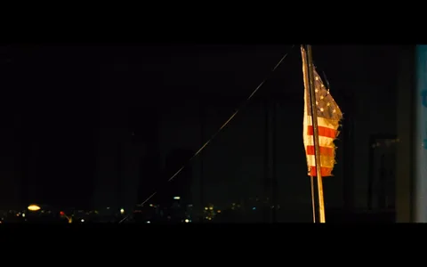 The image depicts an indoor nighttime scene where a U.S. flag is prominently featured. The flag is positioned on the right side, slightly tattered and illuminated by a warm light source. Its colors are slightly muted, but the stars and stripes are clearly visible. The background is dark, suggesting nighttime, with faint city lights visible outside a window, providing a contrast to the flag's brightness. The setting feels quiet and somewhat somber, with the flag standing as the main focal point. There are no people or noticeable activities in the frame, contributing to a still, contemplative atmosphere. This composition evokes a sense of patriotism mixed with nostalgia or reflection. The overall color palette is dark with occasional bright spots from the city lights and the flag's illumination.