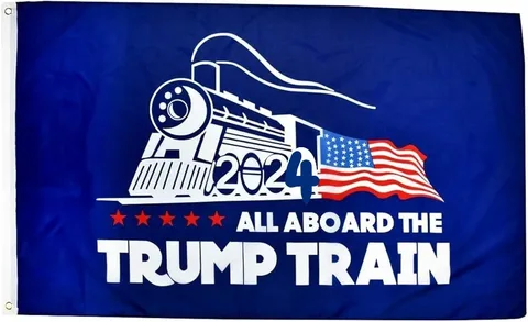 The image features a flag with a political theme. It is predominantly blue and has a stylized white illustration of a train on the left side. The train is facing to the right, and the number "2024" is prominently displayed on its side, indicating a campaign reference for that year. Behind the train, there is an American flag design, adding a patriotic element to the image. Below the train, in bold white text, are the words "ALL ABOARD THE TRUMP TRAIN," which suggest support for Donald Trump's political campaign. The flag includes five red stars, further emphasizing the political theme. The overall color scheme is red, white, and blue, aligning with typical American patriotic colors.