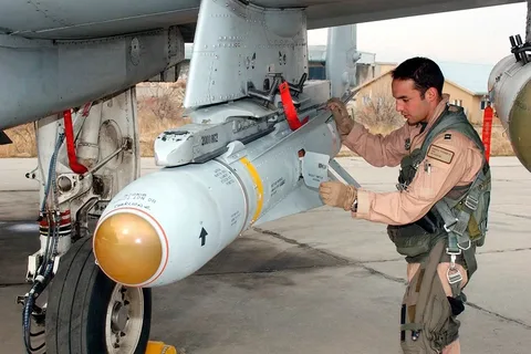 In the image, an individual in a military uniform is conducting an inspection of a missile attached to the undercarriage of an aircraft. The setting appears to be an airbase, with a concrete surface and a building visible in the background. The person is wearing a flight suit in a light brown shade with various badges and patches, along with a harness and gloves, suggesting readiness for operational duties.
The missile is mounted beneath the aircraft, held securely by metal brackets. It has a distinctive design with a white body and a gold-colored tip, featuring various markings and text. Red safety tags hang from the weapon and the aircraft, indicating it is in a non-operational state for inspection. The individual is focused, using one hand to hold a flap open on the missile, likely checking or ensuring a specific condition is met before a flight operation.
The environment is well-lit, likely daytime, providing clear visibility of the details on the missile and the aircraft. The surrounding area is somewhat barren, typical of military installations where operational activities take place. The image captures a moment of routine but critical maintenance, highlighting the precision and attention to detail required in military aviation tasks.