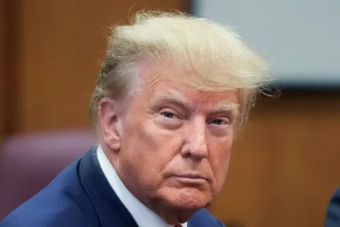 Donald Trump is captured in this image with a serious expression, sitting indoors. His attire includes a suit with a white shirt and red tie, indicative of a formal setting. His hair, a notable feature, is styled in his recognizable fashion. The background is softly blurred, suggesting a focus on him as the subject. The lighting is warm, highlighting his facial features and giving the scene a composed tone. The image seems to be taken in a professional or official environment, providing a sense of gravity and formality. The overall composition centers Donald Trump, making him the focal point, and the close-up framing emphasizes his expression and demeanor.