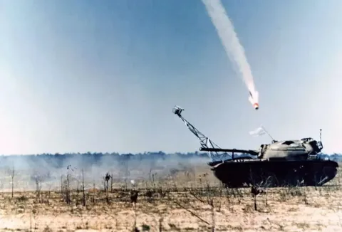 The image captures a dramatic outdoor scene featuring a tank positioned in a barren, desert-like landscape. The tank, with a camouflage pattern, is equipped with a distinctive arm extension mounted on its turret. From this arm, a missile appears to have been launched, with a visible trail of smoke against the clear blue sky. The missile is in mid-flight, heading upward at a steep angle, suggesting it has just been fired. The tank has a white flag attached to it, possibly indicating a training exercise or test rather than active combat. The foreground is sparse, with dry bushes scattered across the sandy ground. The scene is bathed in bright daylight, casting clear shadows and highlighting the movements and mechanisms involved in the missile launch. The setting suggests a military exercise area, emphasizing the technology and precision of modern warfare equipment. The overall mood is one of tension and anticipation, capturing a moment of action frozen in time.