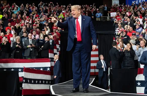 Donald Trump is pictured in this energetic scene, captured at a political rally. He stands confidently on a raised platform, wearing a dark blue suit with a bright red tie, exuding a commanding presence. The background is filled with a dense crowd of supporters, many of whom are capturing the moment on their phones. The atmosphere is lively and vibrant, with attendees wearing red hats and holding signs, some of which bear the phrase "KEEP AMERICA GREAT!"
The setting is an indoor arena, adorned with American flag-themed decorations, adding a patriotic feel to the event. Donald Trump gestures animatedly, pointing into the crowd, suggesting engagement and interaction with the audience. Security personnel are visible nearby, maintaining order as he moves along the stage. The lighting is bright, highlighting Trump and enhancing the visibility of the crowd's enthusiastic expressions. This image vividly conveys the spirited ambiance typical of a political rally.