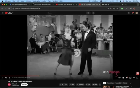 The image is a black-and-white video frame from a YouTube video titled "Top 10 Classic I Love Lucy Moments," published by the channel MsMojo. The scene features a lively dance performance from the classic television show "I Love Lucy." In the foreground, a woman in a sequined dress performs energetically, holding a hat, while a man in a tuxedo stands beside her, watching her dance. The setting appears to be a club or entertainment venue, with an audience seated at tables in the background and a band visible, adding to the lively atmosphere of the scene. The image captures the dynamic motion of the dance, with the woman's movement creating a sense of excitement and entertainment, characteristic of the iconic show. The MsMojo logo is visible in the lower right corner, indicating the publisher of the video.