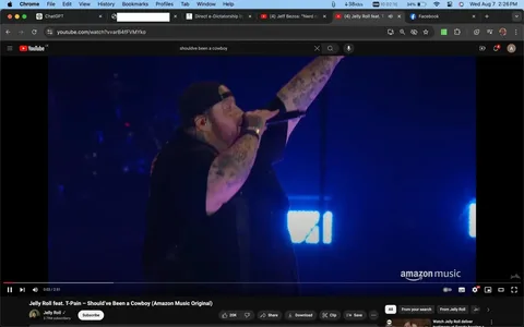 The image shows a live performance screenshot from a YouTube video titled "Jelly Roll feat. T-Pain – Should’ve Been a Cowboy (Amazon Music Original)." The setting is a concert stage, illuminated with blue lighting that enhances the vibrant atmosphere. A performer, identified through the hint as Jelly Roll, is captured mid-performance, holding a microphone in his right hand while raising his left arm enthusiastically toward the audience. He is wearing a black cap backward and a black t-shirt, and his arms are covered in tattoos, adding to his distinctive stage presence.
The YouTube interface is visible, indicating the video length of 2:51 minutes and showing that the video has 20,000 likes. The bottom right corner displays the Amazon Music logo, indicating the collaboration. The channel, Jelly Roll, boasts 3.79 million subscribers, highlighting its popularity. The screenshot captures the energy and engagement of a live musical performance, with Jelly Roll actively connecting with the audience. The user interface includes typical YouTube features such as like, share, and save options.
- Title: "Jelly Roll feat. T-Pain – Should’ve Been a Cowboy (Amazon Music Original)"
- Channel / profile: "Jelly Roll"
- Site / app: "YouTube"
