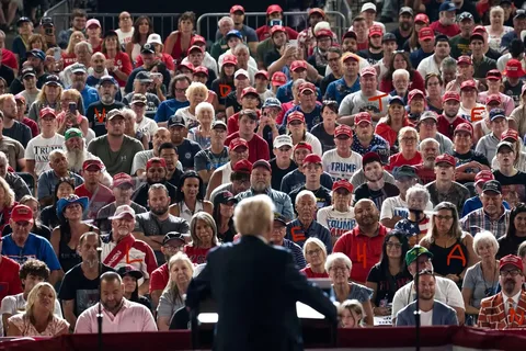 The image shows a large indoor gathering with a crowd of people seated in an auditorium or similar venue. The focus is on the audience, a diverse group clad in various attire, mostly featuring red caps and shirts with slogans or branding. Many caps display the phrase "Make America Great Again," indicative of a political rally. In the foreground, viewed from behind, is a figure at a podium, identified in the hint as Trump. The atmosphere suggests attentiveness as the audience faces the stage, with some capturing the moment using phones. The seating is packed, implying a significant turnout. Lighting is bright, highlighting the gathering's energy and engagement. Visible expressions range from focused listening to enthusiasm. The setup suggests a formal event with organized seating and barriers at the back, containing the crowd's size and scope.