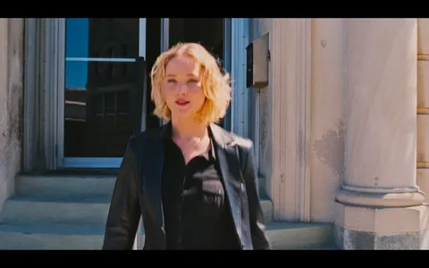 The image shows a person with short blonde hair, standing in front of an entrance to a building. The individual is wearing a black leather jacket over a black shirt, giving a modern and confident appearance. The setting appears to be urban, with the building's stone facade and columns visible. The entrance features glass doors that reflect the bright, sunny day outside. The person's expression seems neutral or focused, and the visual composition centers them against the architectural backdrop. The lighting is natural, suggesting it is taken during the daytime. The overall mood of the image is professional and contemporary.