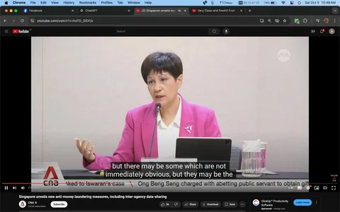 1) Rich description:
The image is a video frame from a YouTube video titled "Singapore unveils new anti-money laundering measures, including inter-agency data-sharing," published by CNA. The scene captures a woman speaking at what appears to be a press conference or formal meeting. She is dressed in a bright pink blazer over a white blouse, adding a formal yet vibrant touch to her attire. Her expression is focused and engaged as she addresses the audience, gesturing with her left hand to emphasize her points. In front of her is a microphone and a tablet, suggesting she is referring to notes or slides. The setting is likely a conference room, with a plain backdrop that keeps the focus on the speaker. The lighting is bright and even, typical of indoor media events. Superimposed text at the bottom provides subtitles for her speech, highlighting the phrase, "but there may be some which are not immediately obvious, but they may be the." The video is part of CNA's coverage on Singapore's new measures against money laundering.
2) On-screen text block:
- Title: "Singapore unveils new anti-money laundering measures, including inter-agency data-sharing"
- Channel / profile: "CNA"
- Site / app: "YouTube"
- Captions / subtitles: "but there may be some which are not immediately obvious, but they may be the"