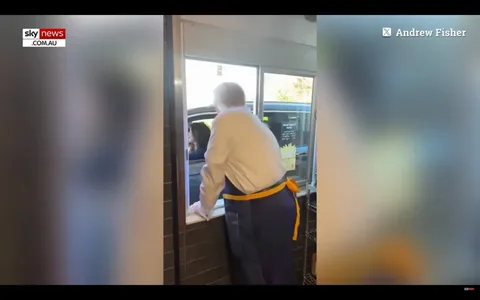 1) Rich description (8–12 sentences): In the image, a man is leaning out of a drive-thru window, engaging with a customer in a vehicle. Based on the hint, this scene includes Donald Trump. The setting is a fast-food restaurant's drive-thru, with an indoor view from behind the counter. The man, wearing a white shirt and a blue apron with a yellow tie, is leaning forward, suggesting an active interaction. The vehicle's driver appears attentive, looking up towards the window. The atmosphere seems casual and dynamic, typical of a drive-thru exchange. The lighting is bright, indicating daytime or a well-lit indoor environment. The video frame includes branding from "Sky News Australia," with the logo visible in the upper left corner. The name "Andrew Fisher" is displayed in the top right, suggesting content attribution.
2) On-screen text block (only include lines that have real text; if a field is unknown or blank, omit that line entirely):
- Site / app: "Sky News Australia"
- Channel / profile: "Andrew Fisher"
- Key OCR Lines:
- "sky news"
- "COM.AU"