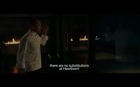 The image is a frame from the film "The Menu." It depicts a tense scene set indoors, possibly in a high-end restaurant. A chef, wearing a white chef's jacket, is prominently featured on the left side of the frame. He appears to be speaking emphatically, gesturing with his hand, which suggests a moment of intensity or confrontation. The lighting is dim, with a warm glow from fires in the background, creating a dramatic and moody atmosphere. A reflection of the chef can be seen on a glass surface, adding depth to the composition. The setting is sophisticated, indicated by the clean lines and the presence of fire, likely from a fireplace or decorative element. The text on the screen reads, "there are no substitutions at Hawthorn!" indicating a strict policy at the restaurant. This scene highlights themes of control and exclusivity typical in high-stakes dining environments as depicted in "The Menu."
- Captions / subtitles: "there are no substitutions at Hawthorn!"
