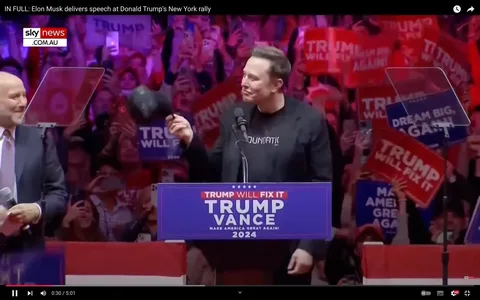 1) Rich description: The image captures a lively scene at a political rally, with Elon Musk standing at a podium. The podium is adorned with a sign reading "TRUMP WILL FIX IT," followed by "TRUMP VANCE" and "MAKE AMERICA GREAT AGAIN! 2024." Elon Musk is wearing a black jacket over a shirt with visible text, gesturing and holding a hat in one hand. The background is filled with a vibrant, cheering crowd holding signs that say "TRUMP WILL FIX IT" and "MAKE AMERICA GREAT AGAIN!" The atmosphere is energetic, with bright purple and red lighting casting a colorful hue over the crowd. The setting suggests a significant political event, with clear branding and messaging aimed at promoting a political campaign. A man in a suit stands nearby, appearing to be engaged in the proceedings, while the audience captures the moment on their smartphones.
2) On-screen text block:
- Title: "IN FULL: Elon Musk delivers speech at Donald Trump's New York rally"
- Channel / profile: "sky news"
- Site / app: "YouTube"