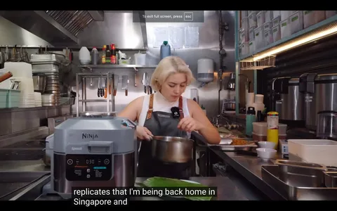 The image depicts a kitchen scene, likely from a cooking video or show. A person with light-colored hair is in focus, wearing a white top and a dark apron. They are engaged in preparing food, holding a pot while standing at a countertop. The kitchen is equipped with various tools and appliances; a Ninja Speedi appliance is prominently displayed in the foreground. In the background, there are shelves with jars, utensils hanging on hooks, and bottles of sauces. The overall atmosphere is busy and professional, indicative of a commercial kitchen. The lighting is bright, highlighting the stainless steel surfaces and various kitchen items.
- Captions / subtitles: "replicates that I'm being back home in Singapore and"