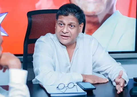Dilith Jayaweera is seen in this image engaging in a conversation. He is sitting at a table, wearing a white shirt that contributes to a professional and approachable appearance. His expression is attentive and engaged, indicating that he is deeply involved in the discussion. The setting suggests an office or meeting room, characterized by a modern black mesh chair and a table cluttered with items such as a notepad, eyeglasses, and a smartphone, suggesting a business environment.
The background features a large screen displaying a partial image that adds vibrancy with its bold red color. The lighting in the room is bright and highlights Dilith Jayaweera's features, enhancing his confident demeanor. His hand gestures further emphasize his involvement in the conversation, possibly indicating a point he is making. The overall atmosphere is one of focused interaction, indicative of a professional meeting or discussion.