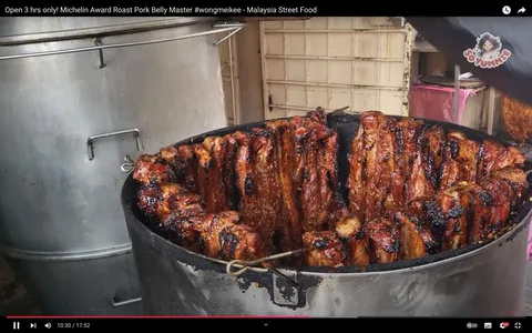 The image showcases a tantalizing display of roast Pork Belly, cooked to perfection in a large, cylindrical metal drum. The pork pieces are charred and glistening with a rich mahogany color, indicating a well-caramelized glaze. The drum is positioned outdoors, suggesting a street food setting typical in Malaysia. The background includes a stainless steel pot and part of a wall, hinting at a casual, bustling environment. The meat is arranged around the circumference of the drum, with visible hooks holding them in place, showing a traditional method of roasting. The atmosphere is likely aromatic, filled with the irresistible scent of spiced meat. This scene is part of a video titled "Open 3 hrs only! Michelin Award Roast Pork Belly Master #wongmeikee - Malaysia Street Food," featured on the channel "Soyummy." The focus is on the culinary expertise and the street food culture, highlighting a unique gastronomic experience in Malaysia.
- Title: "Open 3 hrs only! Michelin Award Roast Pork Belly Master #wongmeikee - Malaysia Street Food"
- Channel / profile: "Soyummy"