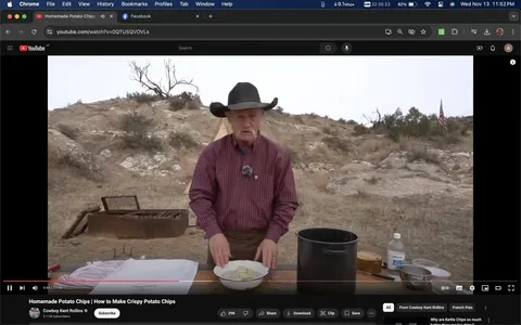 The image is a screenshot from a YouTube video titled "Homemade Potato Chips | How to Make Crispy Potato Chips," presented by Cowboy Kent Rollins. The setting is outdoors in a rural area with dry, rocky terrain prominently visible in the background. Cowboy Kent Rollins is standing behind a wooden table, focusing on the preparation of potato chips. He is wearing a cowboy hat and a red checkered shirt, evoking a rustic, country vibe. Various cooking items are laid out on the table, including a bowl of sliced potatoes, a large pot, and other utensils, suggesting an outdoor cooking setup. An American flag is visible in the background, adding to the countryside atmosphere. The mood is instructional, as Kent appears engaged in demonstrating the recipe. The lighting is natural, consistent with an open, outdoor environment.

- Title: "Homemade Potato Chips | How to Make Crispy Potato Chips"
- Channel / profile: "Cowboy Kent Rollins"
- Site / app: "YouTube"