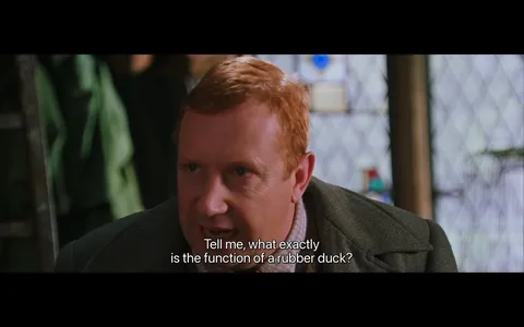 In this scene from the "Harry Potter" series, a character is shown in close-up view, engaging in a conversation indoors. The character has a focused expression and is leaning slightly forward, suggesting curiosity or puzzlement. He has reddish hair and is dressed in a brown tweed suit, adding to the sense of a casual or academic setting. The backdrop features a diamond-patterned window and some indistinct objects, contributing to a cozy, somewhat cluttered atmosphere typical of wizarding environments. The lighting is soft, enhancing the intimate mood of the exchange. The caption "Tell me, what exactly is the function of a rubber duck?" adds a humorous element, as it implies a lack of understanding of everyday Muggle items. This reflects the whimsical and often comedic contrasts between the magical and non-magical worlds in the series.