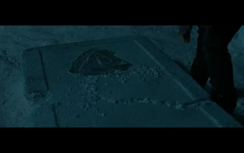 The image depicts a stone slab resting on a sandy or dusty surface, illuminated in a dim light that suggests a nighttime setting. The slab features a distinct symbol, which appears to be a triangle with a circle and a line through it, possibly representing a significant emblem. The surrounding area is marked with fine particles, indicating that the slab has been disturbed or is in a remote location. There are no visible people or additional objects in the frame, emphasizing the focus on the slab and its engraving. The overall atmosphere is mysterious and somewhat eerie, suggesting a scene of discovery or exploration.