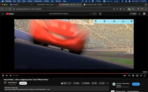 The image appears to be a still from a video featuring an animated scene, likely from the movie "Cars." In the foreground, a bright red car, resembling a race car, is depicted in motion, slightly blurred to indicate speed. The car has a distinctive design with a prominent front grille and large headlights. 

In the background, there is a large crowd in a stadium, suggesting a racing event. The stands are filled with numerous spectators, although individual faces are not discernible due to the distance and focus on the car. The setting appears to be a racetrack, with a smooth asphalt surface visible beneath the car. 

The video interface at the bottom shows that this is a music video for the song "Life Is a Highway" by Rascal Flatts, with the title and artist's name displayed prominently. The video is currently at 4 minutes and 17 seconds into a total length of 4 minutes and 58 seconds. Below the video, there are various interactive elements such as buttons for subscribing, sharing, and downloading, along with a view count of 57 million and a subscriber count of 1.39 million for the channel.

Overall, the image captures a dynamic moment in an animated racing context, emphasizing speed and excitement, with a vibrant color palette dominated by the red of the car against the muted tones of the crowd and stadium.