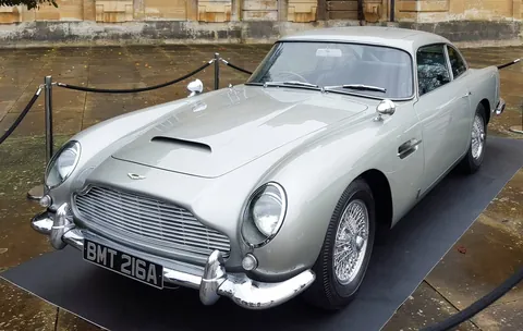 The image features a classic car, specifically a silver Aston Martin, prominently displayed on a black mat. The car has a sleek, aerodynamic design typical of mid-20th century sports cars. Its body is painted in a light silver color, which reflects the surrounding environment, indicating a glossy finish. 

The front of the car showcases a distinctive grille with horizontal slats, flanked by round headlights. The headlights are encased in chrome, adding to the car's elegant appearance. Above the grille, there is a subtle hood scoop, suggesting a performance-oriented design. The license plate reads "BMT 216A," which is a notable detail.

The car's wheels are wire-spoked, contributing to its vintage aesthetic. The tires appear to be black and are mounted on the wheels, which are also shiny and chrome-finished. 

Surrounding the car, there are stanchions with black ropes, indicating that the vehicle is part of an exhibition or display, likely to prevent visitors from getting too close. The background features a stone wall with a textured surface, possibly part of a historic building, and there are some leaves scattered on the ground, suggesting it may be autumn or that it has recently rained, as evidenced by the wet surface of the car.

Overall, the setting is outdoors, with a well-maintained area that appears to be a public space, likely for events or exhibitions. The car is the focal point of the image, showcasing its classic design and craftsmanship.