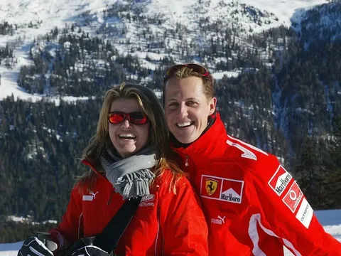 The image features two individuals standing together in a snowy mountainous landscape. Both are smiling and appear to be enjoying their time outdoors.

**Clothing and Appearance:**
- The person on the left has long, wavy hair and is wearing red sunglasses. They are dressed in a bright red jacket with white accents and a logo on the left side, possibly from a brand like Puma. Underneath, they have a gray scarf wrapped around their neck. Their gloves are black, and they are holding something in their left hand, possibly ski equipment.
- The person on the right has short hair and is also wearing red sunglasses. They are dressed in a similar bright red jacket, which features logos, including one that resembles the Ferrari logo and another that appears to be Marlboro. Their jacket has white stripes and additional branding on the sleeves. They are smiling broadly, showing their teeth.

**Background:**
- The background consists of a snowy landscape with mountains in the distance. The mountains are partially covered in snow, and there are patches of greenery visible, indicating a mix of winter and possibly early spring conditions. The sky appears clear and bright, suggesting a sunny day.

**Overall Setting:**
- The scene conveys a recreational outdoor activity, likely skiing or snowboarding, given the attire and the snowy environment. The individuals appear to be in a relaxed and joyful mood, enjoying their time in the mountains.

No additional text, logos, or signs are visible in the image aside from the clothing brands mentioned. The focus is primarily on the two individuals and their cheerful expressions against the scenic backdrop.