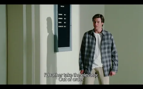 The image depicts a scene in an indoor setting, likely within a building such as an office or a corporate environment. The background features a wall with a directory board mounted on it, which is framed in gray. The directory lists various departments, including "Personnel," "Accounting," "Creative," and "Destructive," all indicating they are located on "INT. 7," suggesting they are on the same floor or level.

In the foreground, a man stands facing the camera. He has short, dark hair styled in a slightly tousled manner. His expression appears to be one of mild surprise or concern, as he looks slightly to his left. He is wearing a plaid shirt that is predominantly gray with darker shades, over a white t-shirt. The shirt is unbuttoned and has long sleeves, which he has rolled up to his elbows. He is also wearing light-colored pants that appear to be casual.

The man is positioned near a wall that has a smooth, light-colored surface, which contrasts with the darker directory board. His shadow is visible on the wall beside him, indicating the direction of the light source. The overall lighting in the scene is bright, suggesting it is daytime.

The text at the bottom of the image reads: "I'd rather take the elevator. Out of order." This text appears to be a subtitle, indicating dialogue from the man in the image. The font is clear and legible, with a white color that stands out against the darker background.

Overall, the setting conveys a typical office environment, with the directory board serving as a functional element for navigation within the building. The man’s attire and expression suggest he is engaged in a conversation or a moment of contemplation regarding the elevator's status.