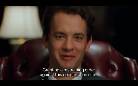 The image features a man seated in a leather chair, likely in an office or formal setting. He has short, dark hair with a slight wave and is wearing a dark suit with a white shirt and a tie. His facial expression is one of slight amusement or confidence, characterized by a subtle smile and relaxed demeanor. The man has light skin and is looking directly at the camera, with his eyes open and engaged.

In the background, there is a soft-focus lamp that emits a warm light, suggesting a cozy or professional atmosphere. The chair he is sitting in has a rich, dark brown leather upholstery, with visible stitching and a tufted design, indicating a classic style.

The text at the bottom of the image reads: "Granting a restraining order against this construction site..." This text appears to be a subtitle, indicating dialogue from a scene, likely from a film or television show. The font is clear and legible, with a white color that contrasts against the darker background.

Overall, the setting conveys a sense of authority and professionalism, likely within a legal or corporate context, given the nature of the dialogue and the man's attire. There are no visible brands, logos, or trademarks in the image. The focus remains on the individual and the text, with no additional objects or products present in the frame.