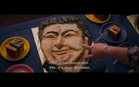 The image depicts a cake designed to resemble a person's face, prominently displayed on a blue tablecloth. The cake features a detailed sculpted face with a smiling expression, characterized by thick, dark hair styled upwards, and pronounced facial features including a broad nose and full cheeks. The cake is decorated with various shades of icing to create depth and texture, particularly around the hair and facial contours.

In the foreground, a hand is pointing at the cake, suggesting interaction or discussion about its likeness. The hand is adorned with a bracelet and appears to belong to a person wearing a floral-patterned sleeve, indicating a casual or festive setting.

Surrounding the cake are several plates with slices of cake, including a rectangular piece with a chocolate layer and a lighter-colored piece, both resting on yellow and blue plates, respectively. The background is blurred, but it suggests an indoor environment, possibly a gathering or celebration, given the presence of food items.

Text at the bottom of the image indicates a conversation, with one person mistakenly identifying the cake as resembling John Goodman, while another corrects them, stating it resembles Alec Baldwin instead. This dialogue adds context to the scene, indicating that the cake is intended to represent a well-known figure, although the specific identity is not confirmed.

Overall, the image captures a lighthearted moment centered around a creatively designed cake, with elements of humor and social interaction evident in the dialogue. The setting appears to be informal, likely at a party or celebration.