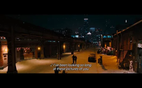 The image depicts a nighttime urban scene, likely set in a city with a skyline featuring tall buildings. The foreground shows a snow-covered street, illuminated by warm yellow lights from street lamps and nearby buildings. The ground is blanketed in snow, creating a serene winter atmosphere. 

On the left side, there is a brick building with a large window displaying colorful holiday lights, suggesting a festive ambiance. The building has a door with a small sign next to it, but the text is not readable. 

In the center of the image, two figures are walking down the street, one appearing to be in a dark coat and the other in a lighter outfit. They are positioned slightly apart, with their backs to the viewer, and seem to be engaged in conversation or simply enjoying the moment. 

To the right, there are parked cars, including a yellow vehicle and a darker one, both partially covered in snow. The street is lined with buildings that have various architectural styles, some featuring brickwork and others with modern glass facades. 

In the background, the skyline is visible with several skyscrapers, some of which have lights illuminating their outlines. Notably, one building has a distinctive spire, and another has a bright, colorful display, possibly an advertisement or decorative lighting. 

The sky is dark with hints of falling snowflakes, adding to the wintery feel of the scene. Overall, the image captures a quiet, picturesque moment in a bustling urban environment during winter.