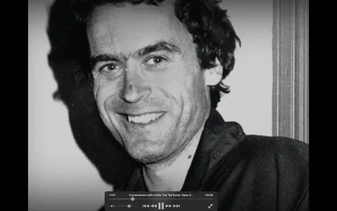 The image features a black-and-white photograph of a man with dark, slightly curly hair and a light complexion. He is smiling broadly, displaying a set of straight teeth. His facial expression appears friendly and approachable. The man has prominent eyebrows and a slight stubble on his face. 

He is wearing a dark-colored jacket, which is partially visible in the image. The collar of the jacket is turned down, revealing a light-colored shirt underneath. The background is plain and light, possibly white, which contrasts with his darker clothing. There are no visible logos, text, or other identifiable features in the background.

The lighting in the image creates a soft shadow on the left side of his face, enhancing the contours of his features. The photograph appears to be a close-up, focusing primarily on his face and upper torso. The overall composition is centered on the man, with no other objects or people present in the frame. 

The image does not contain any readable text, headlines, or signs, and there are no visible brands or products. The setting does not provide specific context, as it appears to be a simple portrait without any identifiable location or event indicators.