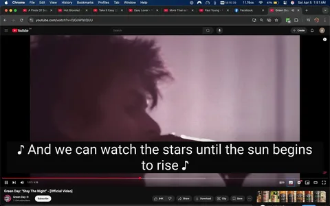 The image shows a scene from a YouTube video titled "Stay The Night" by Green Day, indicated by the logo and channel name at the bottom. The video is playing, and the timestamp shows it is at 1 minute and 57 seconds out of a total of 4 minutes and 38 seconds. 

In the foreground, there is a close-up of a person, likely a musician, who appears to be singing or performing. The individual has short, tousled hair and is partially turned to the side, with their mouth near a microphone. The background is blurred, featuring soft, muted colors, predominantly shades of pink and purple, which create a dreamy atmosphere.

At the bottom of the image, there are subtitles displayed in white text against a black background, reading: "And we can watch the stars until the sun begins to rise." The text is large and clear, making it easy to read.

The YouTube interface is visible at the top, showing various tabs and bookmarks, indicating that the user is on a computer. The video controls, including options to like, share, download, and clip, are also visible beneath the video. The screen shows a collection of beverage cans at the bottom, suggesting a casual viewing environment.

Overall, the image captures a moment from a music video, focusing on the performance aspect while providing context through the YouTube interface.