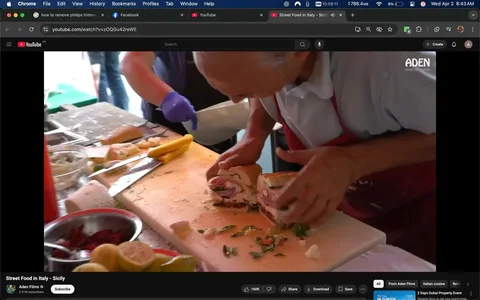 The image depicts a food preparation scene, likely in a market or street food setting. In the foreground, an elderly man is engaged in assembling a sandwich. He has gray hair and is wearing a white shirt with a red apron. His expression is focused as he carefully holds a sandwich that appears to be filled with various meats and possibly vegetables. The sandwich is cut in half, revealing layers of what looks like ham, salami, and other ingredients.

The man’s right hand is positioned on the sandwich, while his left hand is partially visible, possibly holding a knife or another ingredient. He is working on a white cutting board that has remnants of food, including bits of herbs and crumbs, indicating recent activity.

In the background, another person is visible, wearing a blue glove and a shirt, possibly assisting in the food preparation. This person is out of focus but appears to be reaching for something off-screen.

The setting includes various kitchen tools and ingredients scattered across the countertop. There are bowls containing what looks like chopped vegetables or condiments, and a few lemons and limes are visible, suggesting the use of fresh ingredients. A knife is also present on the cutting board, alongside a yellow object that could be a banana or another type of food.

The overall environment appears to be lively and bustling, typical of a street food market, with bright lighting and a casual atmosphere. The background is slightly blurred, focusing attention on the man and his task. There are no visible signs, logos, or text in the image that indicate specific brands or products.