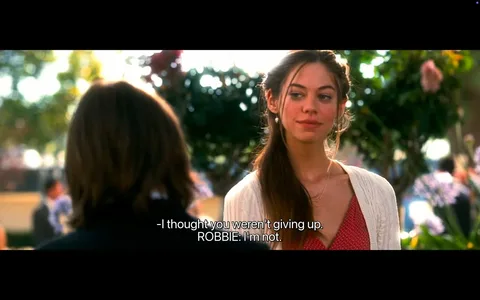 The image depicts a scene from a film, featuring two characters engaged in conversation. 

**Text:**
At the bottom of the image, there are subtitles that read:
- "-I thought you weren't giving up."
- "ROBBIE: I'm not."

**Characters:**
1. **Female Character:**
   - She has long, brown hair styled with a loose braid.
   - Her facial expression appears thoughtful or slightly amused.
   - She is wearing a light-colored, knitted cardigan over a red dress with a pattern.
   - She has a delicate necklace visible around her neck.

2. **Male Character:**
   - The male character is partially visible, with his back facing the camera.
   - He has medium-length, dark hair.
   - He is dressed in a dark suit, suggesting a formal or semi-formal occasion.

**Setting:**
- The background features a lush, green environment with trees and flowering plants, indicating an outdoor setting, possibly a park or garden.
- There are blurred figures in the background, suggesting other people are present, but they are not clearly defined.
- The lighting appears warm and bright, indicating a sunny day.

**Overall Context:**
The scene captures a moment of dialogue between the two characters, set against a vibrant and lively backdrop, which may suggest a social gathering or significant event. The focus is on their interaction, with the female character looking directly at the male character, who is turned slightly away.