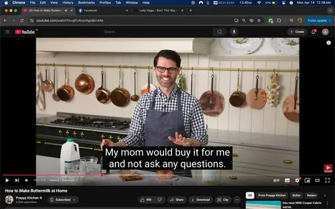 The image shows a kitchen setting where a person is presenting a cooking demonstration. The individual is wearing a checkered shirt, with the colors being predominantly white and dark blue, and an apron that is dark gray. They have glasses and a friendly smile, indicating a welcoming demeanor. The person is gesturing with their hands, possibly explaining a point or engaging with an audience.

In the background, there are several copper pots and pans hanging from a brass rod, which adds a classic touch to the kitchen decor. The kitchen features a large stove with multiple burners, and the cabinetry is a light color, likely white or cream. There are decorative plates displayed on the wall above the stove, contributing to the aesthetic of the kitchen.

On the countertop, there are various items visible: a clear glass measuring cup, a bottle, and a carton of milk with a green cap, indicating it may be whole milk. There is also a bowl that appears to contain a mixture, possibly for the recipe being discussed. The text overlay on the video reads, "My mom would buy it for me and not ask any questions," suggesting a light-hearted or nostalgic commentary related to the cooking process.

The overall atmosphere of the kitchen is bright and inviting, with natural light illuminating the space. The setting appears to be a home kitchen, designed for cooking demonstrations or tutorials.