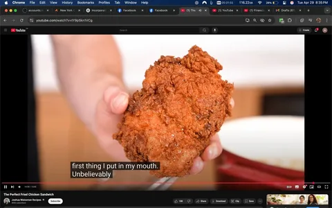 The image shows a close-up of a hand holding a piece of fried chicken. The chicken appears golden brown and crispy, with a textured surface that suggests a crunchy coating. The piece is large, indicating it may be a thigh or breast portion. The hand holding the chicken is partially visible, with fingers gripping the piece firmly. 

In the background, there is a blurred kitchen setting, with a light-colored surface that seems to be a countertop. A white bowl is positioned in the foreground, partially out of focus, suggesting it may contain additional food or ingredients. 

At the bottom of the image, there are subtitles that read: "first thing I put in my mouth. Unbelievably." This text is displayed in a black box with white lettering, indicating it is part of a video or cooking show. 

The overall composition emphasizes the fried chicken, making it the focal point of the image. The lighting appears bright and inviting, enhancing the appeal of the food. There are no visible logos, brands, or identifiable people in the image. The setting suggests a casual cooking environment, likely within a home or a cooking studio.