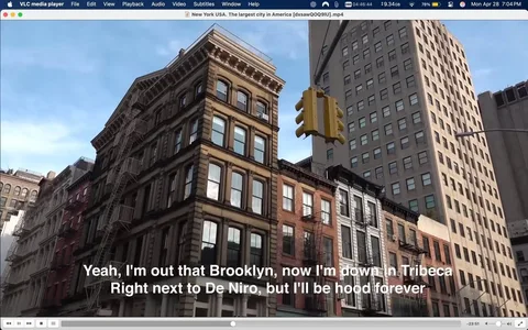 The image shows a street scene in New York City, specifically in the Tribeca neighborhood. The foreground features a row of historic buildings with a mix of architectural styles, including brownstone and brick facades. The buildings have multiple stories, with some displaying fire escapes and large windows. The leftmost building has a reddish hue, while the adjacent buildings are more neutral in color, with browns and creams.

In the center of the image, there is a yellow traffic light hanging from a pole, indicating a pedestrian crossing. The light is positioned above the street, which is not fully visible in the image. The sky is partly cloudy, with blue patches visible, suggesting a clear day.

In the background, taller modern buildings rise, contrasting with the older structures in the foreground. These buildings have a more uniform appearance, with many windows and a sleek design. The scene captures a blend of old and new architecture typical of urban environments.

At the bottom of the image, there is visible text in white font that reads: "Yeah, I'm out that Brooklyn, now I'm down in Tribeca Right next to De Niro, but I'll be hood forever." This text appears to be part of a subtitle or caption, likely related to a video or audio content.

Overall, the image conveys a vibrant urban atmosphere, highlighting the architectural diversity of New York City while incorporating elements of contemporary culture through the text.