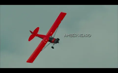The image features a bright red biplane flying at an angle against a cloudy sky. The aircraft has a classic design, characterized by its open cockpit and large wings. The wings are painted in a vibrant red, and the plane appears to be performing an aerial maneuver, possibly a roll or a loop, as it is tilted to one side. 

In the foreground, the name "AMBER HEARD" is displayed prominently in a bold, white font. The text is positioned centrally on the right side of the image, contrasting against the background and ensuring visibility. The font style is clean and modern, contributing to the cinematic feel of the scene.

The background consists of a pale blue sky with scattered clouds, providing a soft contrast to the vivid red of the plane. The overall atmosphere suggests a sense of adventure and excitement, typical of aerial scenes in films.

No other objects, people, or identifiable landmarks are present in the image. The focus remains solely on the biplane and the text associated with it. The image does not contain any logos, trademarks, or additional text beyond the name displayed.