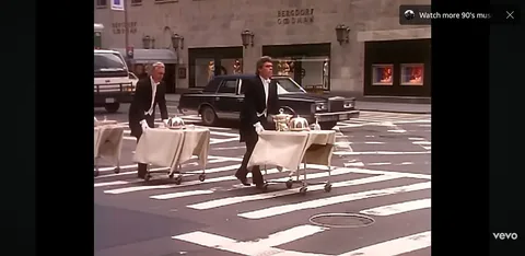 The image depicts two men dressed in formal attire, specifically black tuxedos with white shirts and bow ties, pushing carts across a crosswalk. Each cart is covered with a beige cloth and holds silver serving trays, suggesting they are delivering food or drinks. The man on the left has short, light-colored hair and is wearing white gloves, while the man on the right has darker hair styled in a wavy manner and also wears white gloves. Both men appear focused on their task as they navigate the street.

The background features a city street with a mix of vehicles, including a black sedan and a white truck. The setting appears to be an urban area, likely in a high-end shopping district, as indicated by the presence of a store with the name "Bergdorf Goodman" visible on the building in the background. The architecture is characterized by a stone facade and large windows displaying mannequins and clothing, suggesting a luxury retail environment.

The crosswalk is marked with white stripes, and the scene is bathed in daylight, indicating it is likely daytime. The overall atmosphere seems busy, typical of a metropolitan area, with the men actively engaging in their duties amidst the urban hustle. There are no visible signs, logos, or additional text in the image apart from the store name. The focus remains on the two men and their carts as they traverse the street.