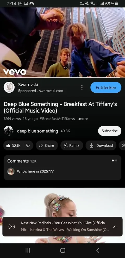 The image displays a mobile screen showing a video playback interface for the song "Breakfast At Tiffany's" by the band Deep Blue Something. The video has accumulated 69 million views and was uploaded 15 years ago. The title of the video is prominently displayed at the top, along with the band name. Below the video, there is a logo for Vevo in the corner, indicating that the video is hosted on that platform.

The interface includes a sponsored section featuring the brand "Swarovski," with a link to their website. Below the video title, there are hashtags, specifically #BreakfastAtTiffanys, which may relate to the song's theme or content. The channel name "deep blue something" is visible, along with a subscriber count of 40.3K.

The interaction buttons below the video include options to like (with 324K likes), share, remix, and download. There is a comment section with a total of 12,000 comments, and one comment reads, "Who's here in 2025???"

In the lower part of the screen, there is a preview for the next video, which is "You Get What You Give" by New Radicals, along with a mix of songs including "Walking On Sunshine" by Katrina & The Waves. The background of the next video preview features a woman adorned with colorful accessories, though her details are not fully visible.

The overall setting appears to be a digital media platform, likely a music streaming service, with a clean and user-friendly interface designed for video playback and interaction. The background is predominantly dark, emphasizing the video content and interface elements.